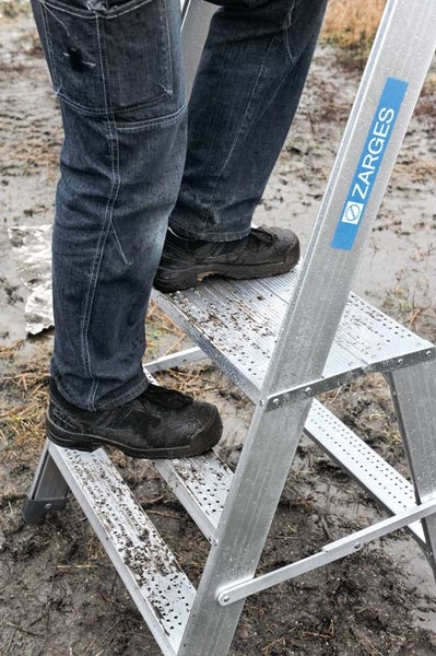 Person steht auf einer Zarges Aluminium Stehleiter mit rutschfesten Stufen auf einem schlammigen Untergrund.