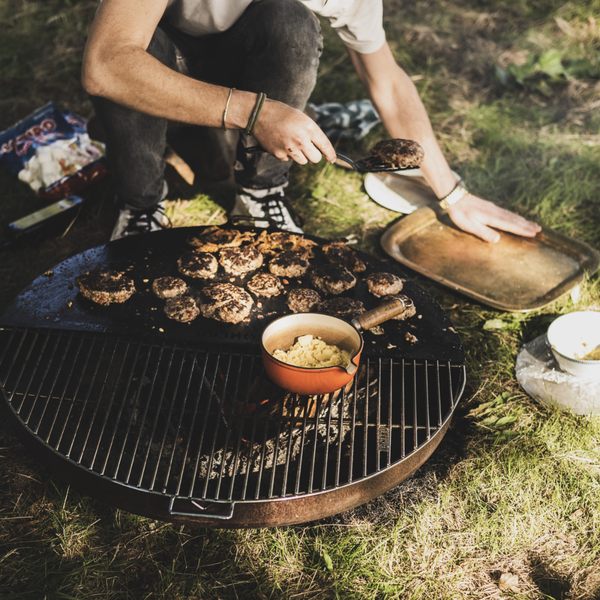 Person grillt Burger auf einer runden Feuerschale mit Grillrost und Plancha-Platte im Freien auf einer Wiese.