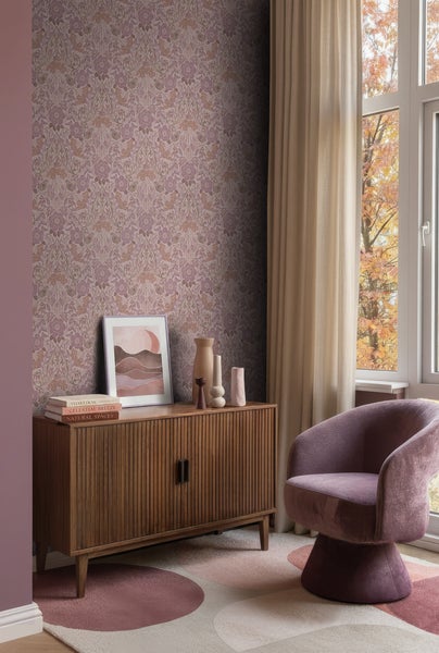 Wohnzimmer mit floral gemusterter Tapete in Mauve, Sideboard aus Holz, lila Sessel und Teppich vor hellem Fenster mit herbstlichem Laub.