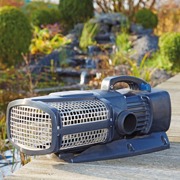 Teichpumpe aus Kunststoff mit Filterkorb und Tragegriff auf einem hellen Holzsteg vor einem bepflanzten Gartenteich mit Bachlauf.