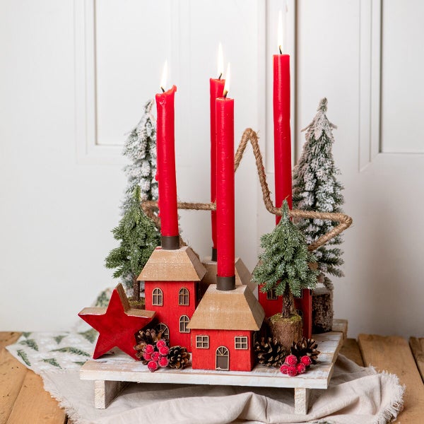 Adventskranz mit vier brennenden Kerzen auf einem Holztablett dekoriert mit kleinen Häusern, Tannenbäumen, Sternen, Zapfen und Beeren.