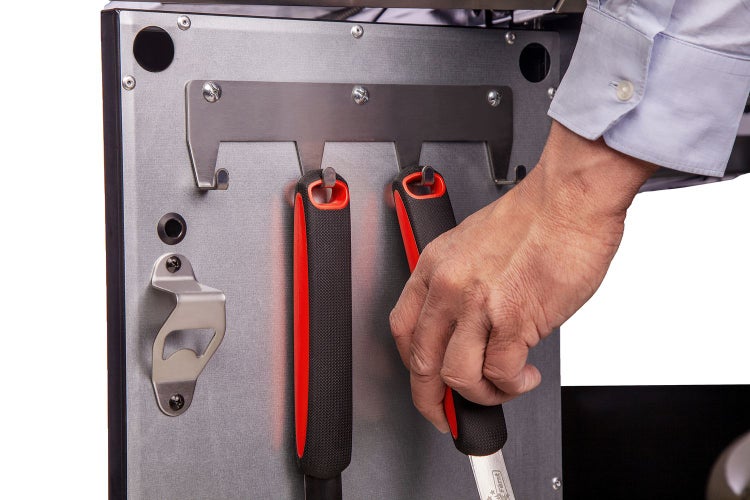 Detailansicht einer Grillstation mit Grillwerkzeugen, die an einer Halterung aufgehängt sind, sowie einem Flaschenöffner