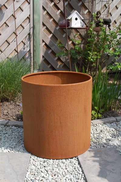 Zylindrischer Pflanzkübel aus Metall in Edelrost-Optik in einem hellen Garten auf Kies und Steinplatten vor einem Holzlattenzaun.