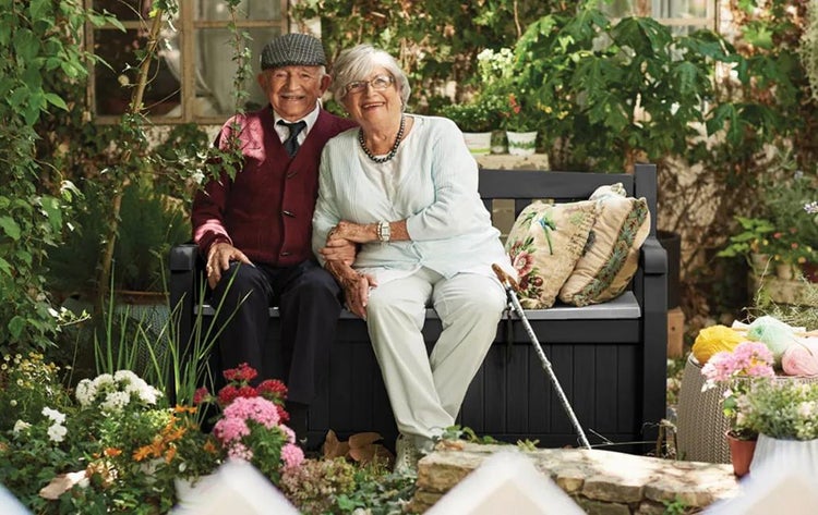 Ein älteres Ehepaar sitzt auf einer anthrazitfarbenen Gartenbank aus Kunststoff mit integrierter Sitztruhe in einem begrünten Garten mit Blumen.