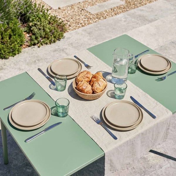 Gedeckter Gartentisch im Freien mit grüner Tischplatte, beigem Geschirr, Gläsern, Besteck und einem Korb mit Croissants auf einer Terrasse.