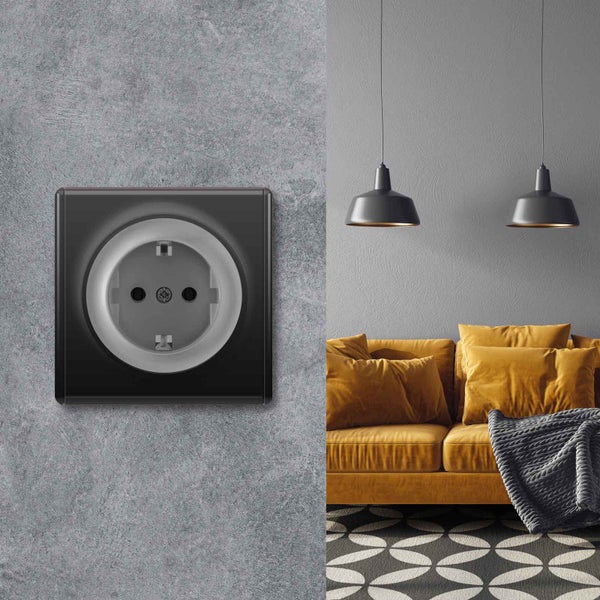 Steckdose in modernem Design an einer Wand mit Betonoptik neben einem Wohnzimmer mit Sofa und Hängeleuchten