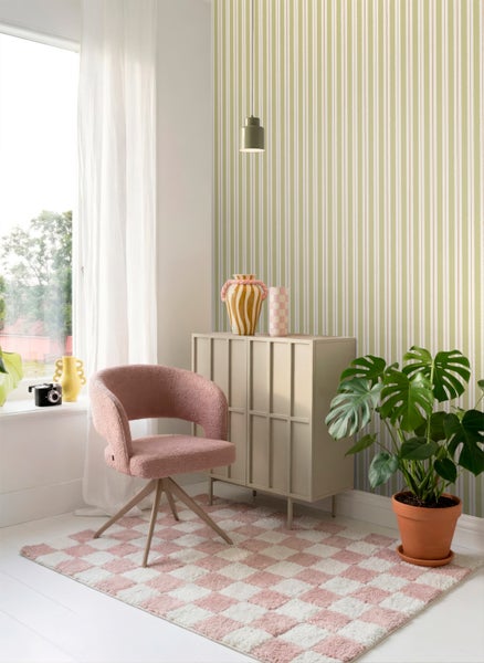 Heller Raum mit grün-cremefarben gestreifter Tapete, rosa Polstersessel, hellbeigem Schrank und einer großen Monstera-Pflanze auf einem karierten Teppich.