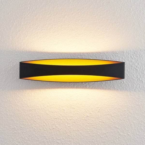 Wandleuchte in geschwungener Form, schwarz mit goldfarbener Innenseite, Lichtstrahl nach oben und unten an einer strukturierten Wand.