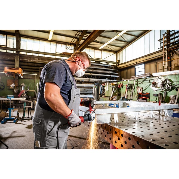 Handwerker mit Schutzkleidung bearbeitet ein Metallteil mit einem Winkelschleifer an einer Werkbank in einer Industriehalle