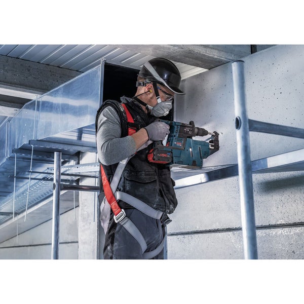 Ein Mann bohrt mit einem Akku-Bohrhammer ein Loch in eine Wand. Er trägt eine Schutzbrille, einen Helm und eine Atemschutzmaske.