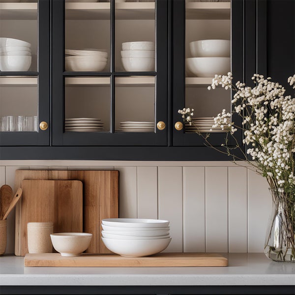 Organisierte Küche mit schwarzen Hängeschränken mit Glasfront, weißen Keramikschalen, Holzschneidebrettern, hellen vertikalen Fliesen und Blumenvase.
