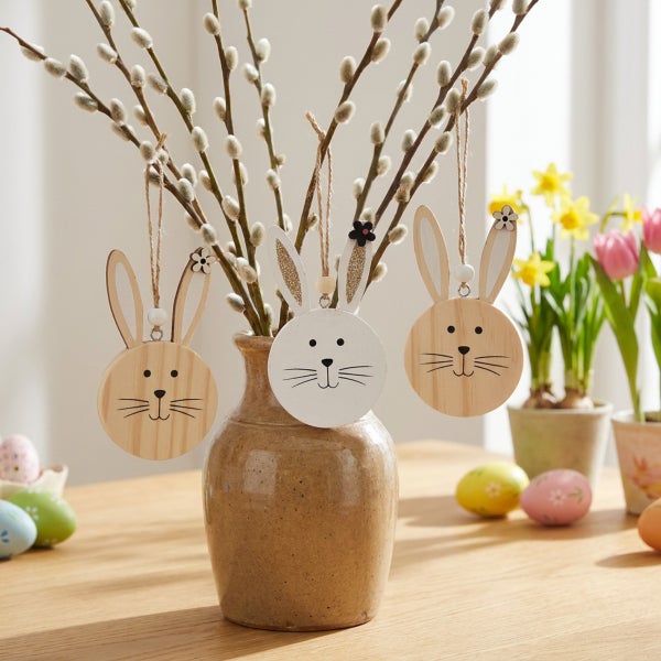 Drei Osteranhänger in Hasenform aus Holz hängen an Weidenkätzchenzweigen in einer Tonvase als österliche Tischdekoration.