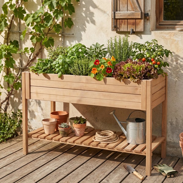 Hochbeet aus Holz bepflanzt mit Kräutern und Gemüse, inklusive Ablagefach mit Tontöpfen und Gießkanne auf einer Holzterrasse.