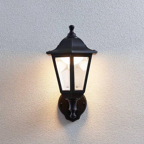 Klassische Außenwandleuchte in schwarzer Laternenform aus Metall und Glas, eingeschaltet an einer Wand.