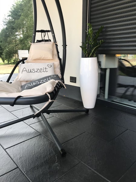 Moderner schwarzer Hängesessel mit Kissen auf einer Terrasse mit dunklen Bodenfliesen in Schieferoptik und einer Pflanze im hohen weißen Topf.