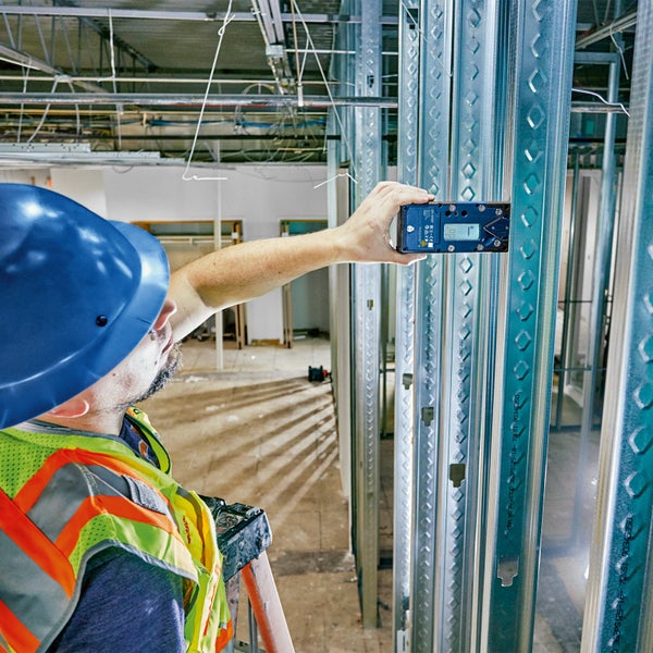 Ein Bauarbeiter mit Schutzhelm und Warnweste benutzt eine digitale Wasserwaage an einer Metallwand auf einer Baustelle.