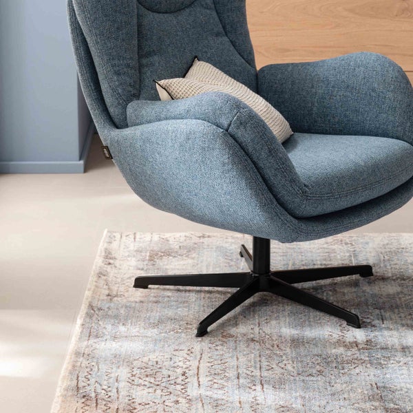 Polstersessel mit blauem Stoffbezug, schwarzem Metalldrehfuß und Kissen auf einem gemusterten Teppich.