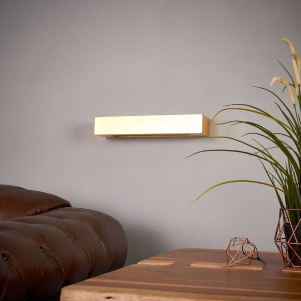 Moderne rechteckige LED-Wandleuchte in Goldoptik in einem hellen, organisierten Wohnzimmer mit Ledersofa, Holztisch und einer Zimmerpflanze.