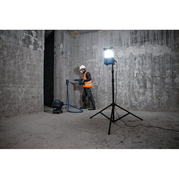 Ein Arbeiter bohrt in einer Baustelle mit einem Bohrhammer und einem Staubsauger neben einer Baustellenlampe.