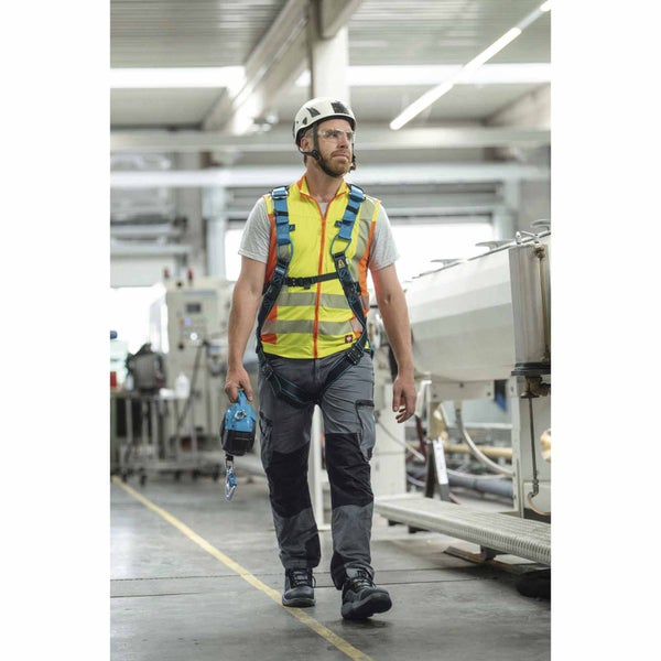 Mann in Industriehalle mit PSA: Auffanggurt, Warnweste, Schutzhelm, Schutzbrille, Arbeitshose und Höhensicherungsgerät.