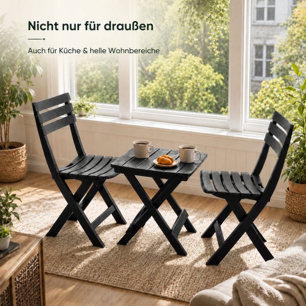 Ein Set aus einem Klapptisch und zwei Klappstühlen aus Holz in einem hellen Wohnbereich vor einem Fenster mit Zimmerpflanzen.