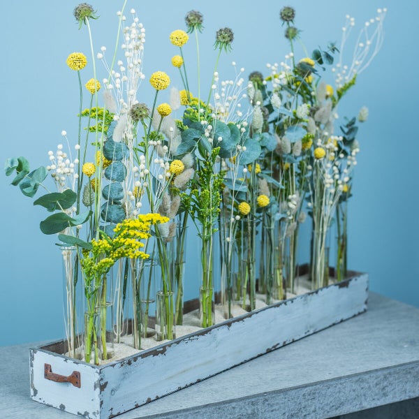 Dekorative Blumenarrangement bestehend aus Trockenblumen in Glasröhrchen auf einem länglichen Holztablett mit rustikaler Oberfläche