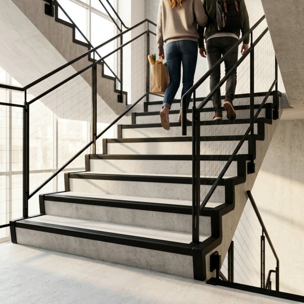 Moderne Betontreppe mit schwarzem Metallgeländer und rutschhemmenden Kantenprofilen im Innenbereich. Zwei Personen steigen die Treppe hinauf.