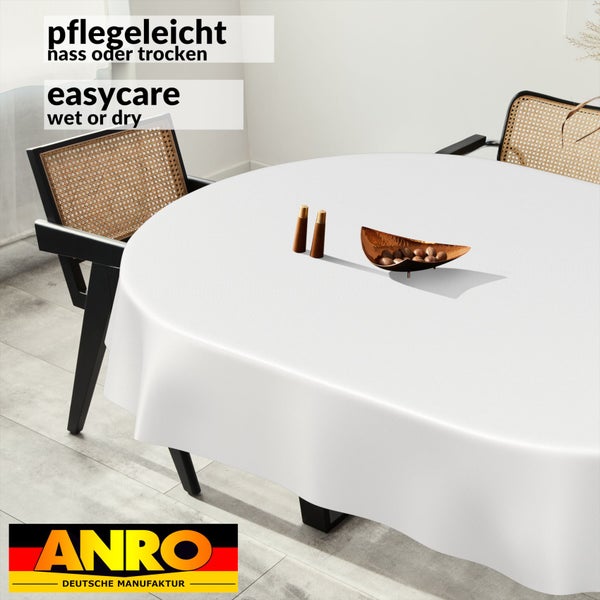 Weiße runde Tischdecke auf einem Esstisch in einem hellen Zimmer mit Rattan-Stühlen. Pflegeleicht nass oder trocken. ANRO Deutsche Manufaktur Logo.