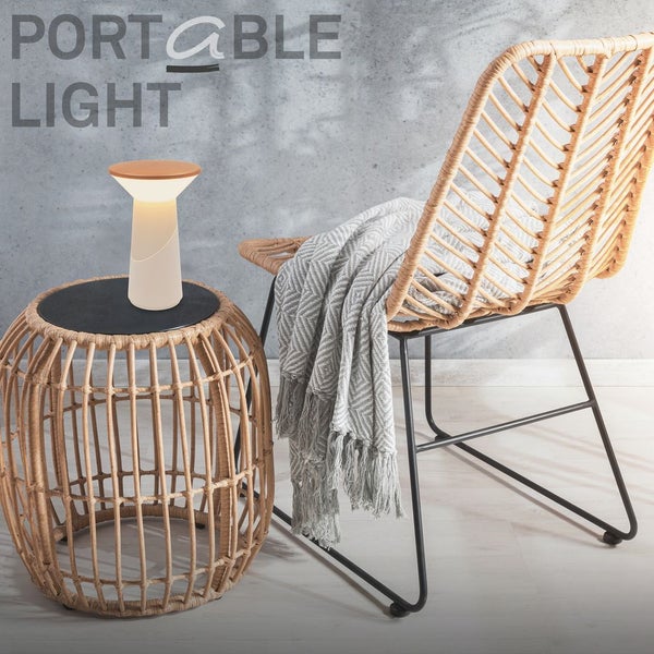 Tragbare LED-Tischleuchte auf einem Beistelltisch aus Rattan neben einem Stuhl aus Rattan mit einer gemusterten Decke vor einer strukturierten Wand.