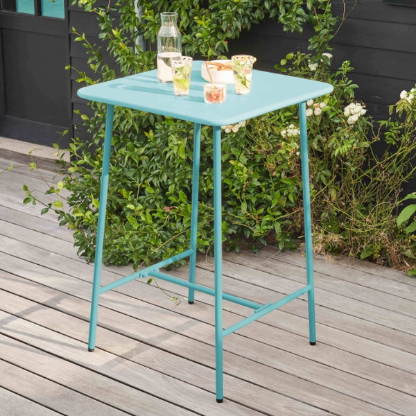 Hoher quadratischer Stehtisch aus Metall in Türkis auf einer Holzterrasse, gedeckt mit Getränken und Snacks, umgeben von Pflanzen.