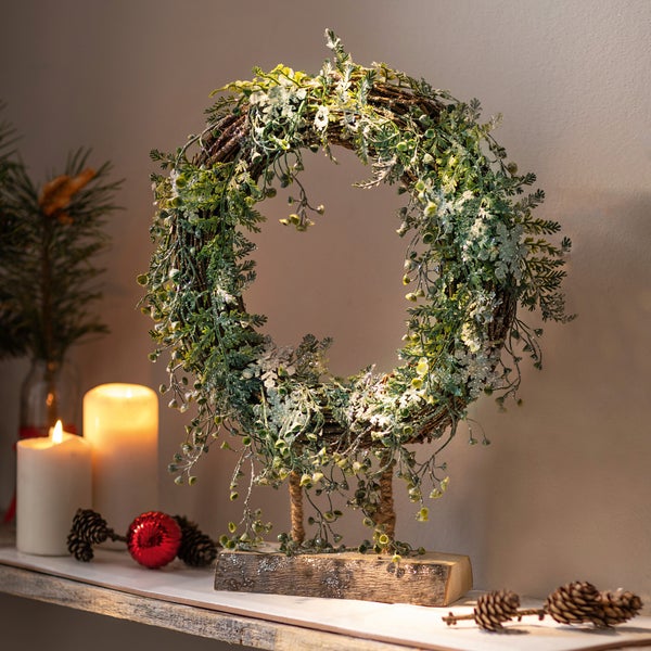 Dekorativer Weihnachtskranz mit Pflanzen und Kunstschnee auf Holzständer.