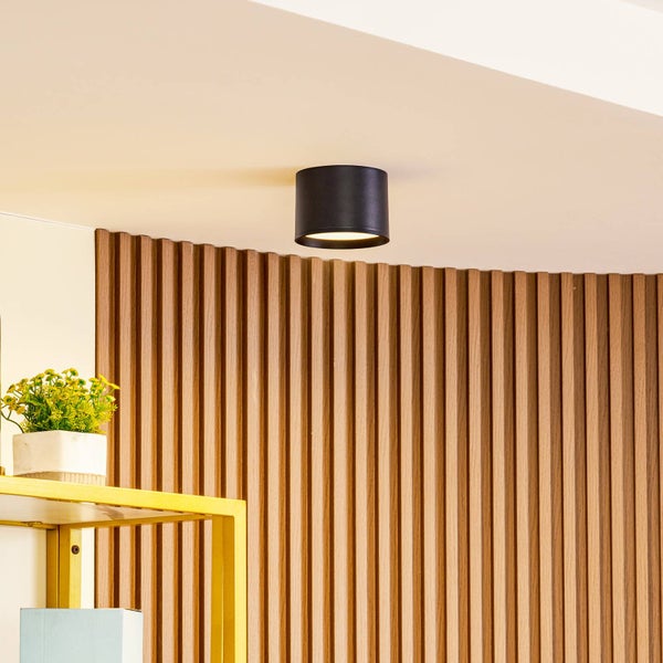 Moderne zylindrische Deckenleuchte in Schwarz, montiert an einer hellen Decke vor einer Wand mit vertikalen Holzlamellen.