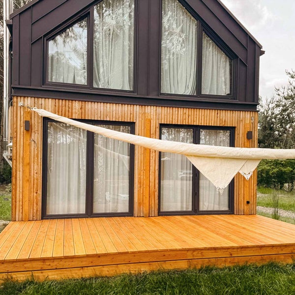 Moderne Hausfassade aus Holz mit großen Fenstern, Holzterrasse und einem hellen Sonnensegel als Schattenspender.