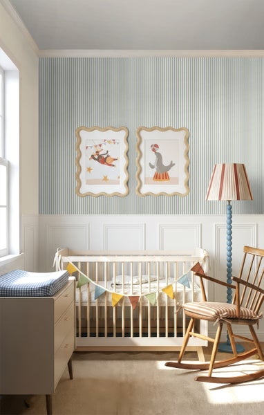 Helles Babyzimmer mit gestreifter Tapete, weißer Wandvertäfelung, hölzernem Gitterbett, Schaukelstuhl und Zirkus-Wandbildern.