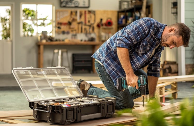 Ein Mann arbeitet mit einem Akkumulator-Bohrschrauber an Holzbrettern in einer hellen Werkstatt neben einem offenen Organizer für Kleinteile.