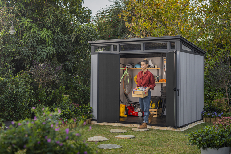 Garten Szene mit Gerätehaus, Rasen und einer Frau, die eine Kiste trägt