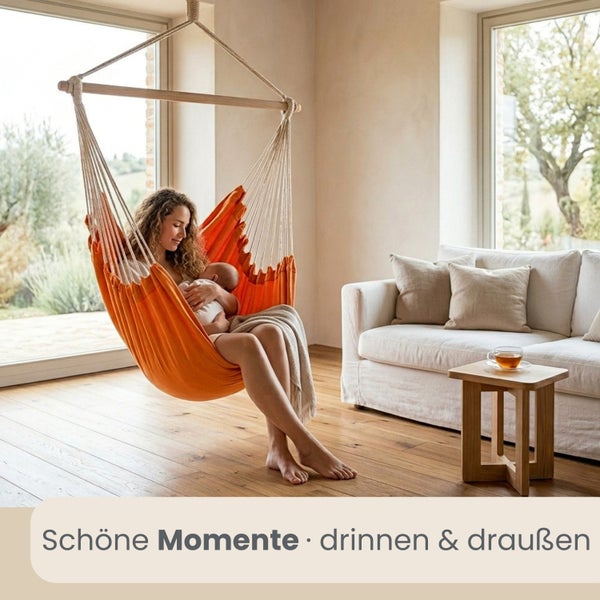 Frau mit Baby in einem orangefarbenen Hängesessel aus Stoff in einem hellen Wohnzimmer mit Holzfußboden und Sofa.
