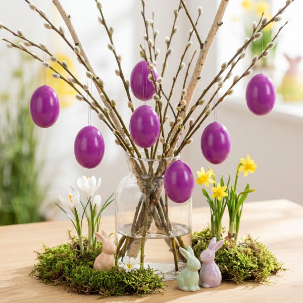 Osterdekoration mit Weidenzweigen in einer Glasvase, geschmückt mit violetten Eiern, Osterhasenfiguren und Frühlingsblumen auf einem Holztisch.