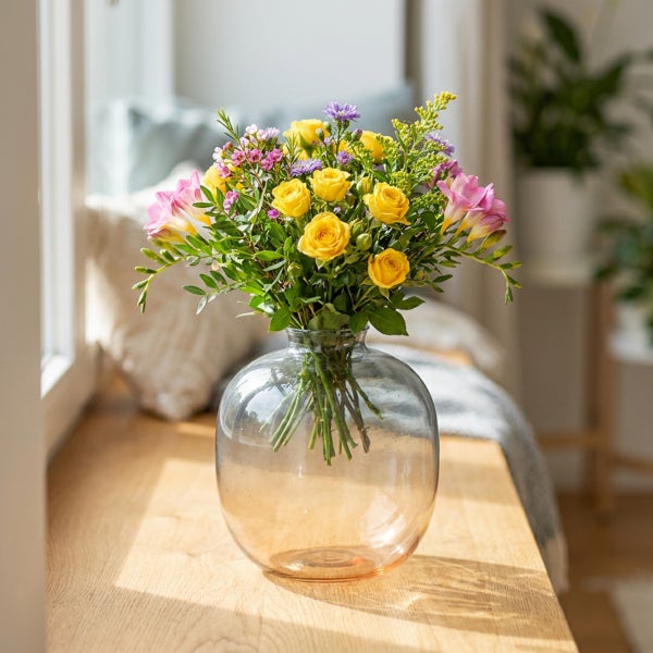 Ein bunter Blumenstrauß aus gelben Rosen und rosa Freesien in einer bauchigen Glasvase auf einer Holzfensterbank.