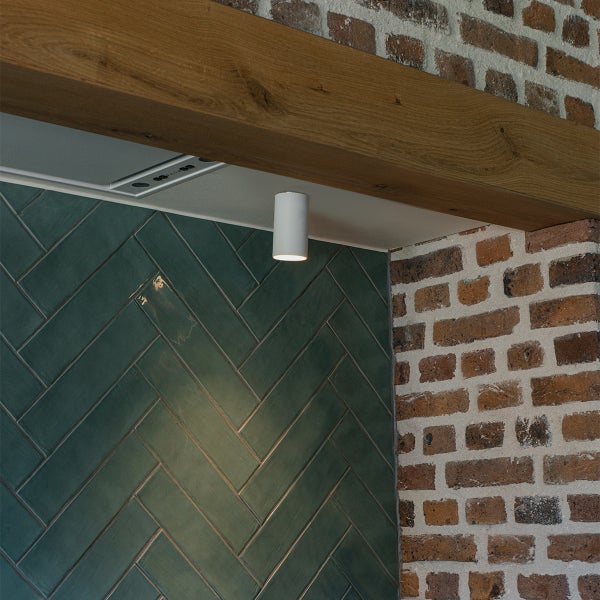 Innenaufnahme mit einer Wand aus grünen Fischgrätenfliesen, einer Ziegelwand und einem Holzrahmen mit einer zylindrischen Deckenleuchte