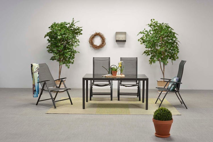 Gartenset bestehend aus schwarzem Gartentisch mit Glasplatte und vier grauen Hochlehner Klappstühlen auf einem Teppich vor zwei künstlichen Bäumen.