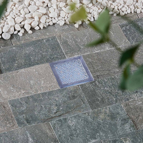 Quadratischer Glasstein eingelassen in eine Terrasse aus Natursteinplatten und Kieselsteinen.