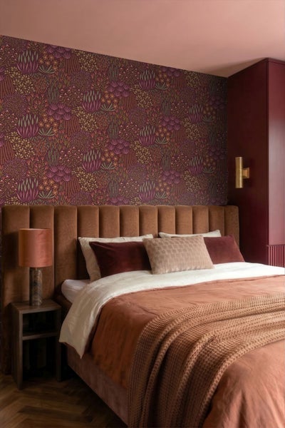 Schlafzimmer mit floral gemusterter Vliestapete, gepolstertem Kopfteil und gemütlichem Bett in warmen Erdtönen.