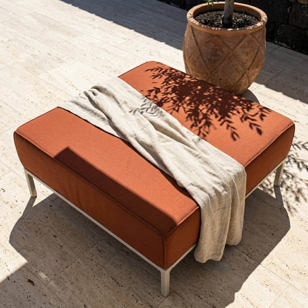 Großer quadratischer Outdoor-Hocker mit terracottafarbenem Polster und hellem Metallgestell, dekoriert mit einer Wohndecke auf einer Steinterrasse.