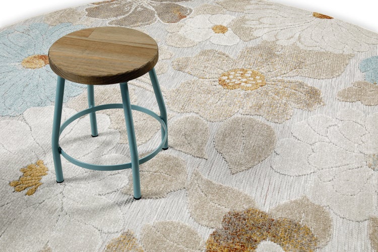 Runder Outdoor-Teppich mit Blumenmuster in Braun, Beige und Blau auf einer Holzterrasse neben einem Sofa und Poufs in heller Umgebung.