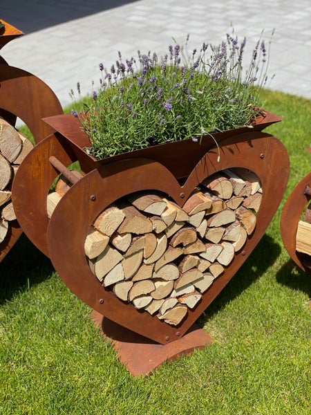 Herzförmiges Kaminholzregal aus Edelrost mit integriertem Pflanzgefäß, bepflanzt mit Lavendel auf einer Rasenfläche.