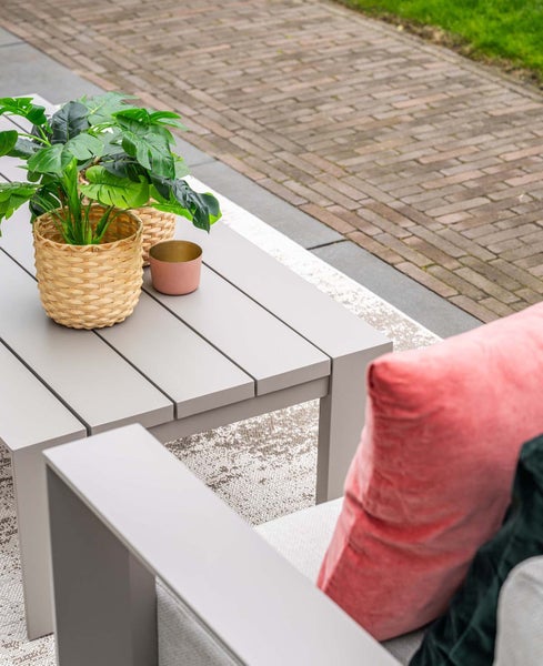 Moderner grauer Gartentisch aus Latten mit geflochtenem Pflanzkorb und rosa Kissen im Außenbereich.