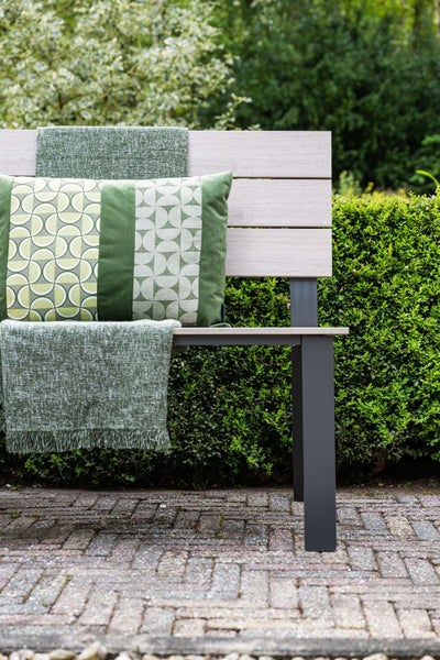 Gartenbank aus Holz mit Metallgestell, dekoriert mit einem Kissen mit geometrischem Muster und einer strukturierten Decke, vor einer Hecke.