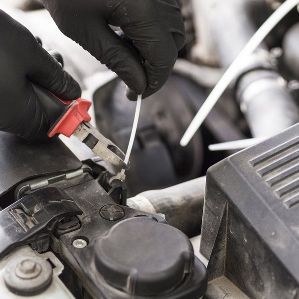 Person mit Handschuhen im Motorraum eines Autos bei der Verwendung eines Seitenschneiders zum Durchtrennen eines Kabelbinders aus Kunststoff.