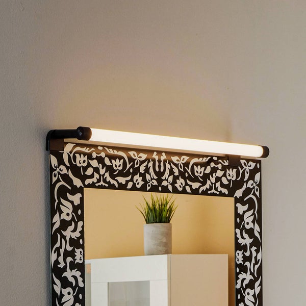 Schwarze LED-Bilderleuchte in zylindrischer Form, die helles Licht über einen verzierten Spiegel mit schwarz-weißem Rahmen wirft.